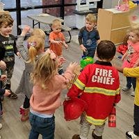 Children playing in a circle, one child wearing a "Fire Chief" jacket