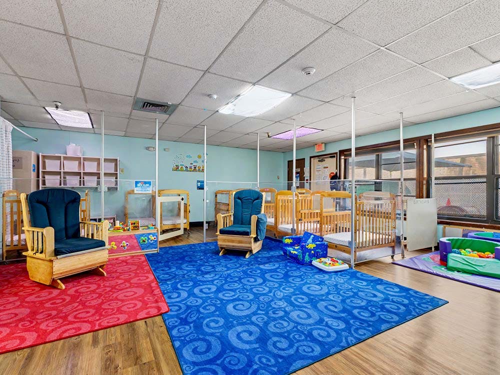 A daycare room with multiple cribs, two blue rocking chairs, and colorful rugs