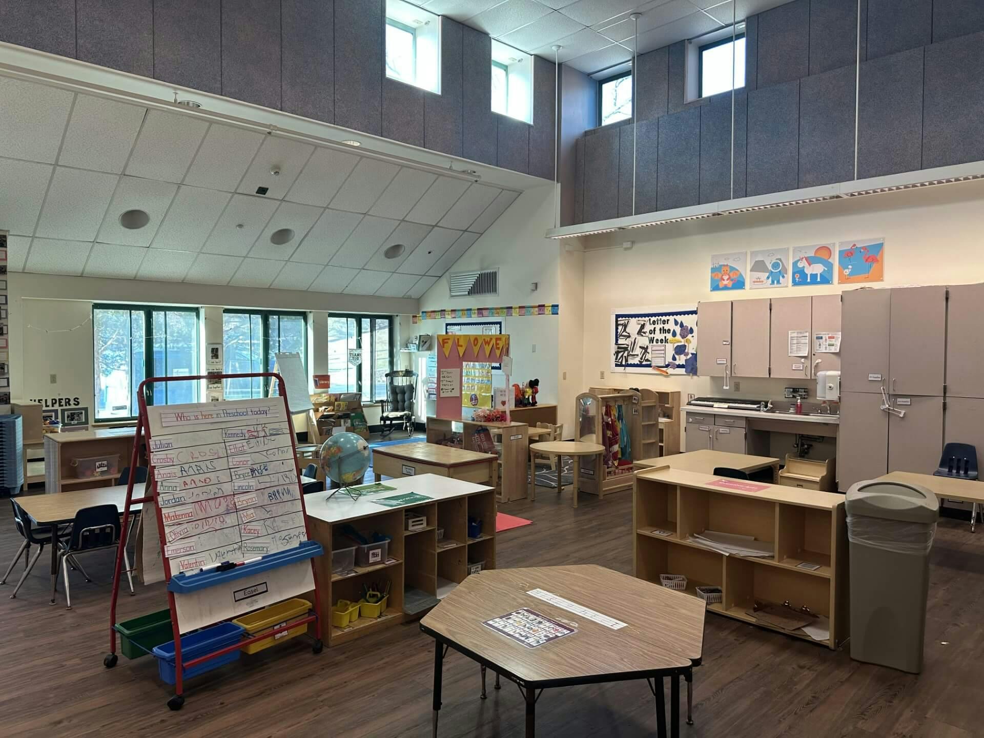 An empty preschool classroom with tables, chairs, and a whiteboard listing names