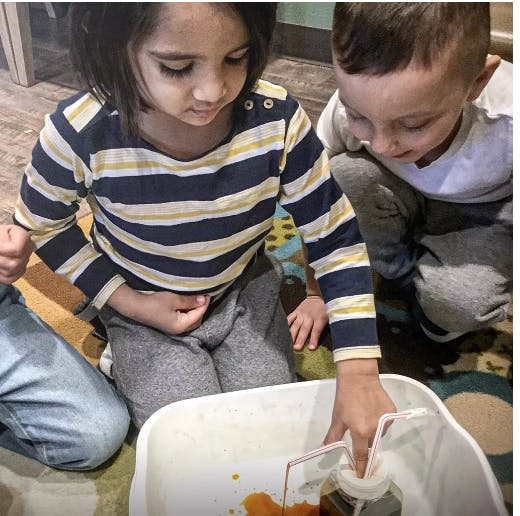 Child in striped shirt reaching into a white bin with another child nearby