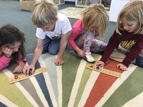 Four children playing with rulers and small objects on a rug