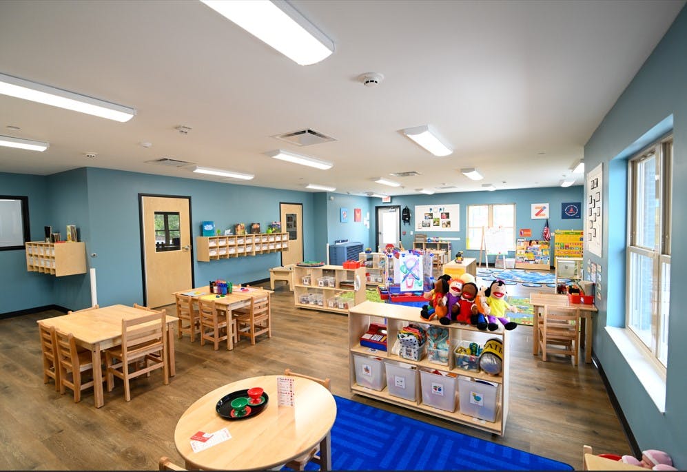 Empty classroom with small wooden tables and chairs, shelves of toys, and blue walls
