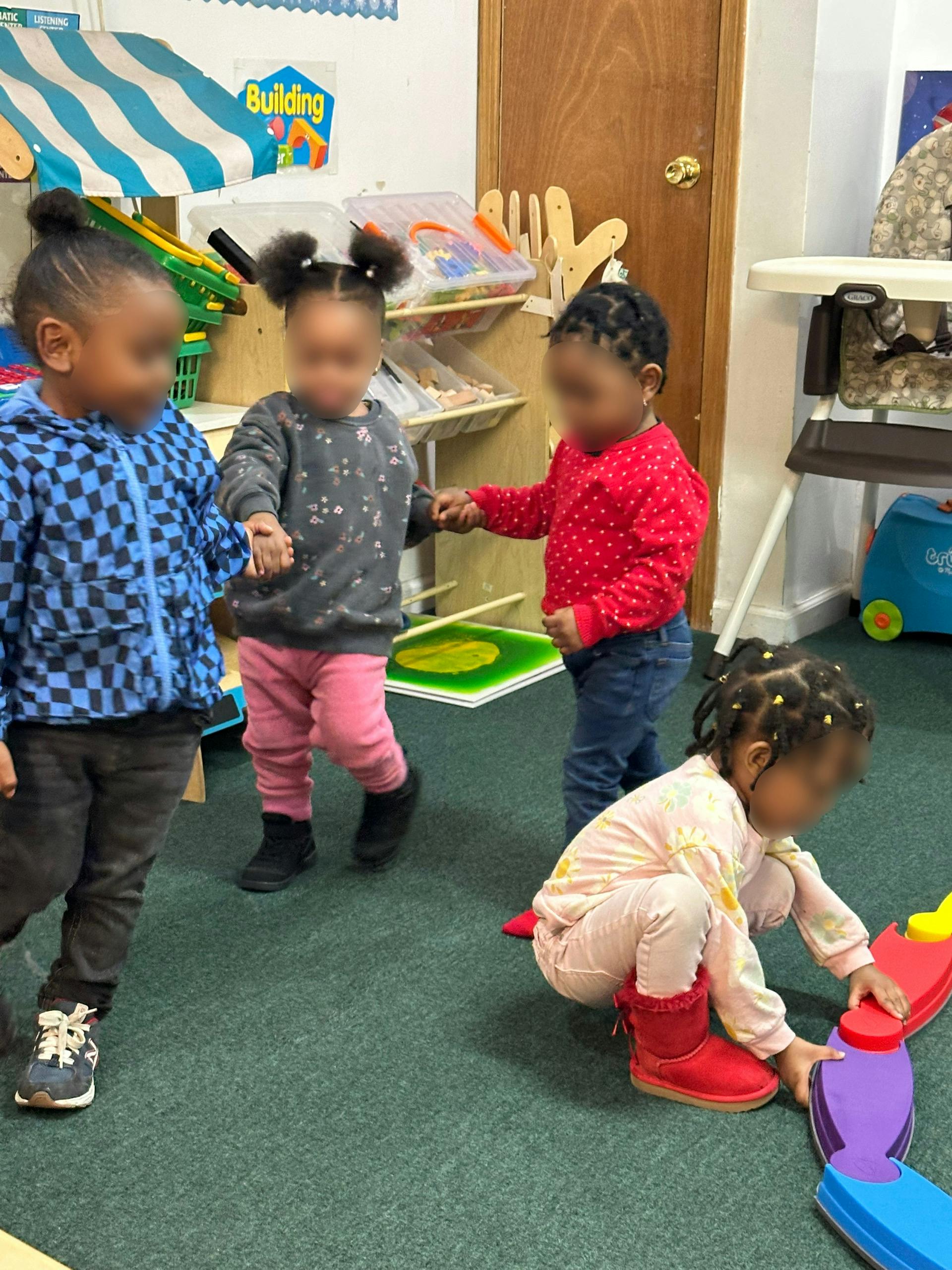 Three children holding hands and one child on the floor playing with toys in a classroom
