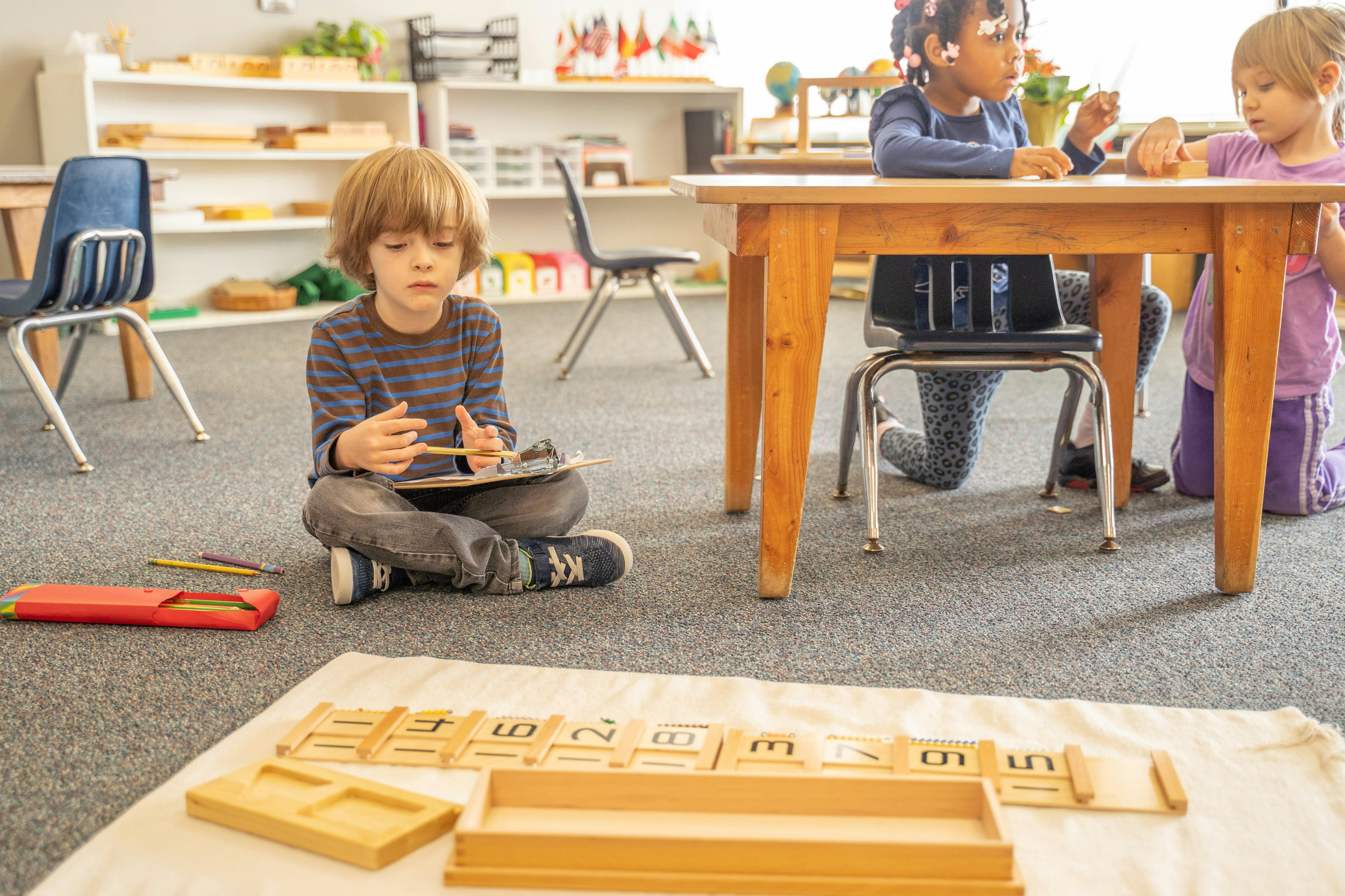 Child sitting on floor with tablet, other children at table in classroom