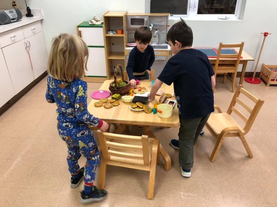 Three children playing with toy food at a small table in a classroom