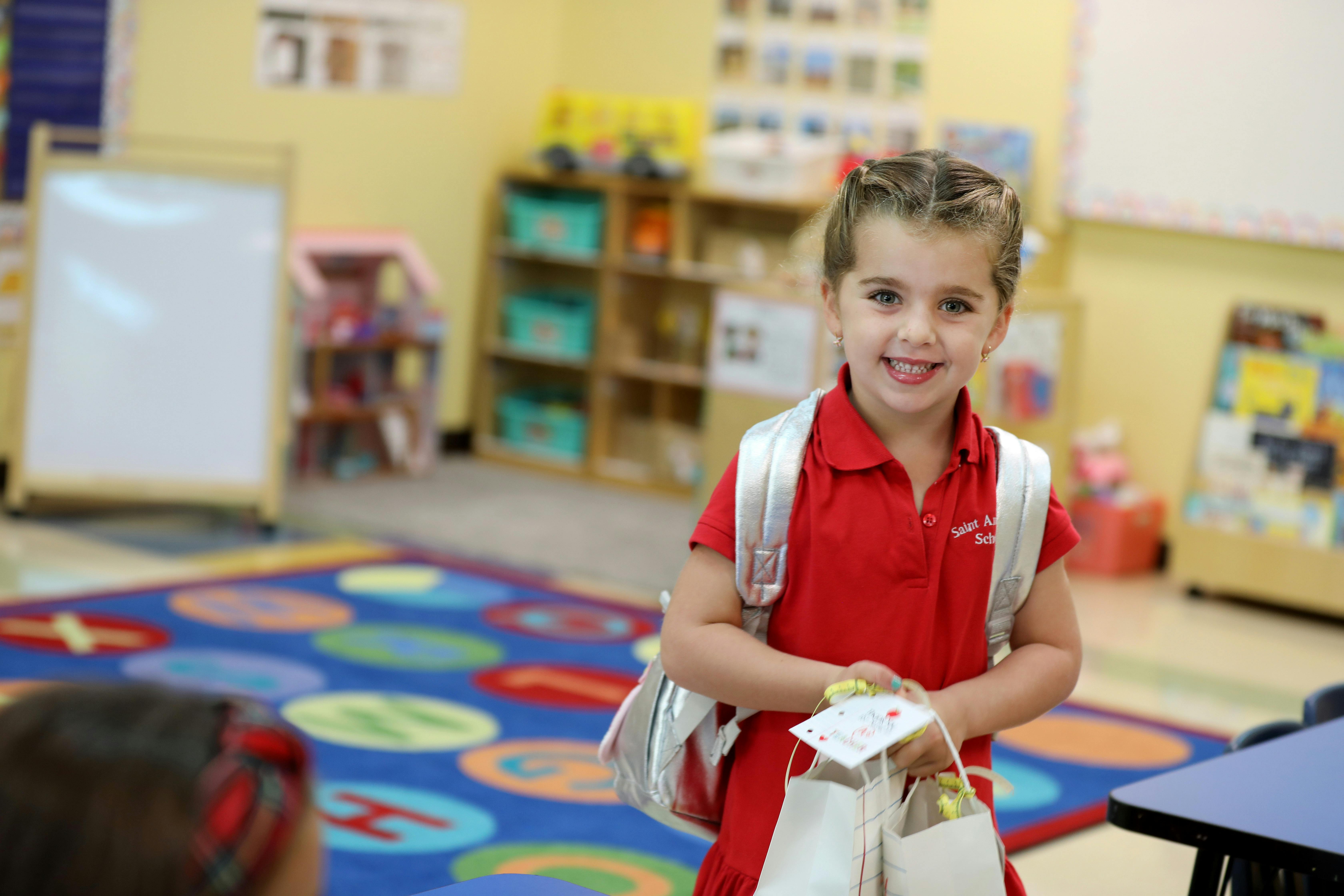 Child in red uniform and silver backpack holding gift bags in a classroom