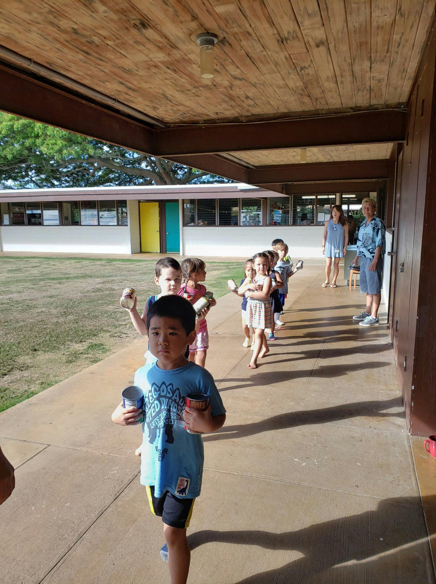 Children in a line on a covered walkway, some holding cups
