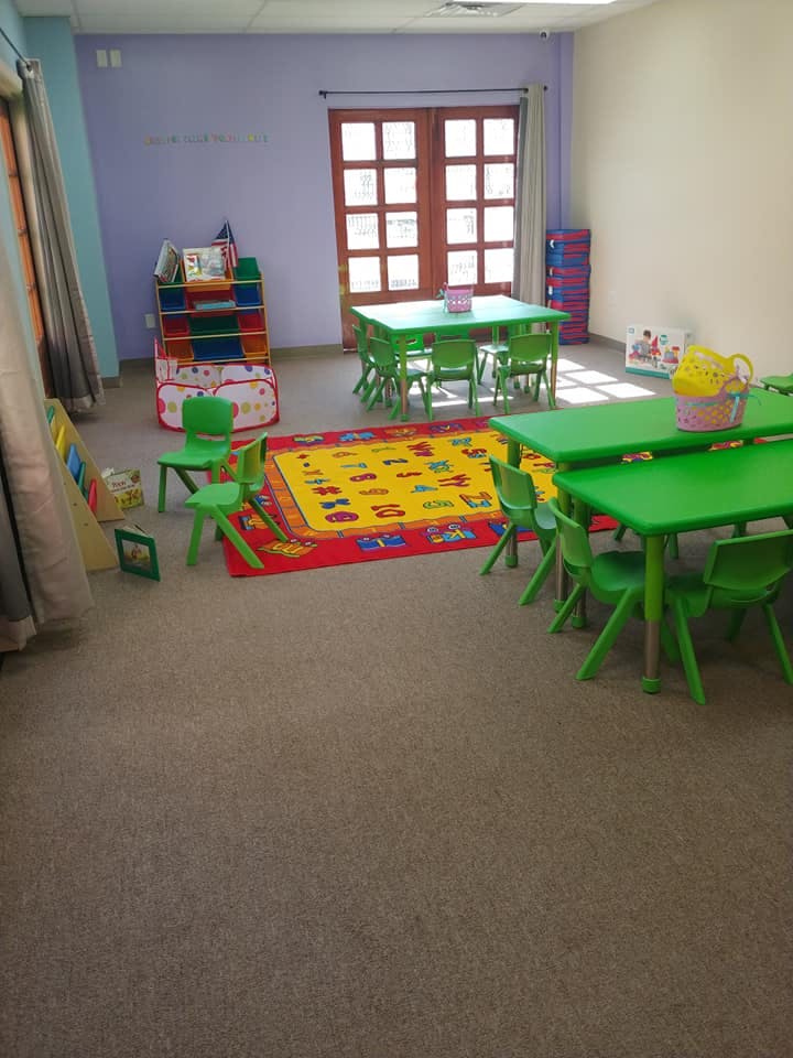 Classroom with green tables and chairs, colorful rug, and storage shelves