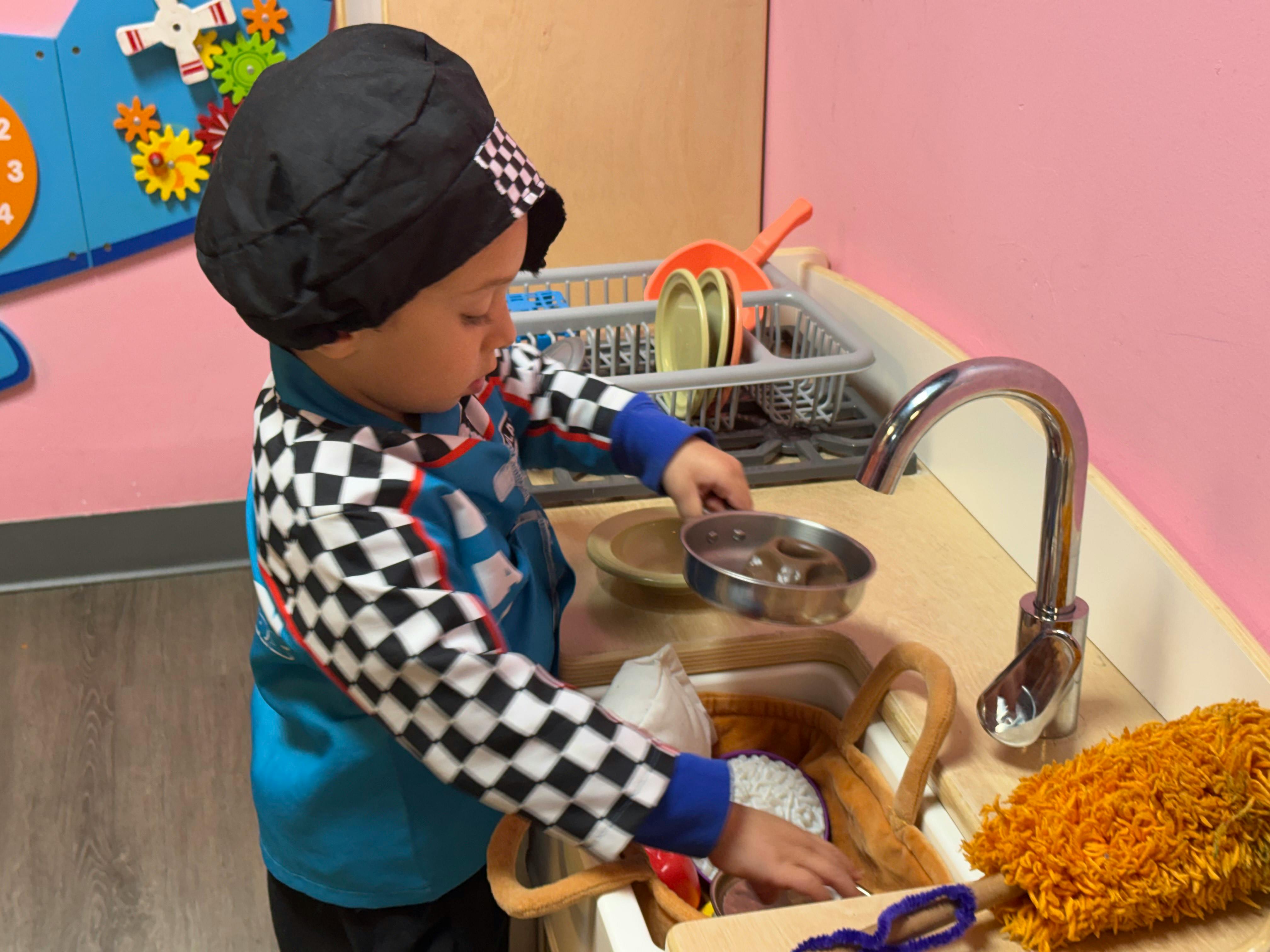 Child playing at a toy kitchen sink, holding a small pot