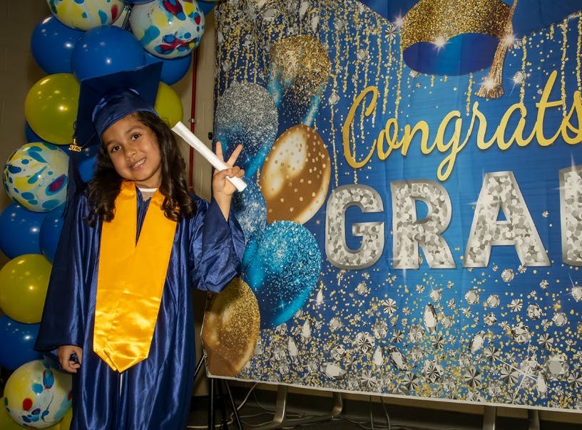 Child in graduation cap and gown holding a diploma, making a peace sign