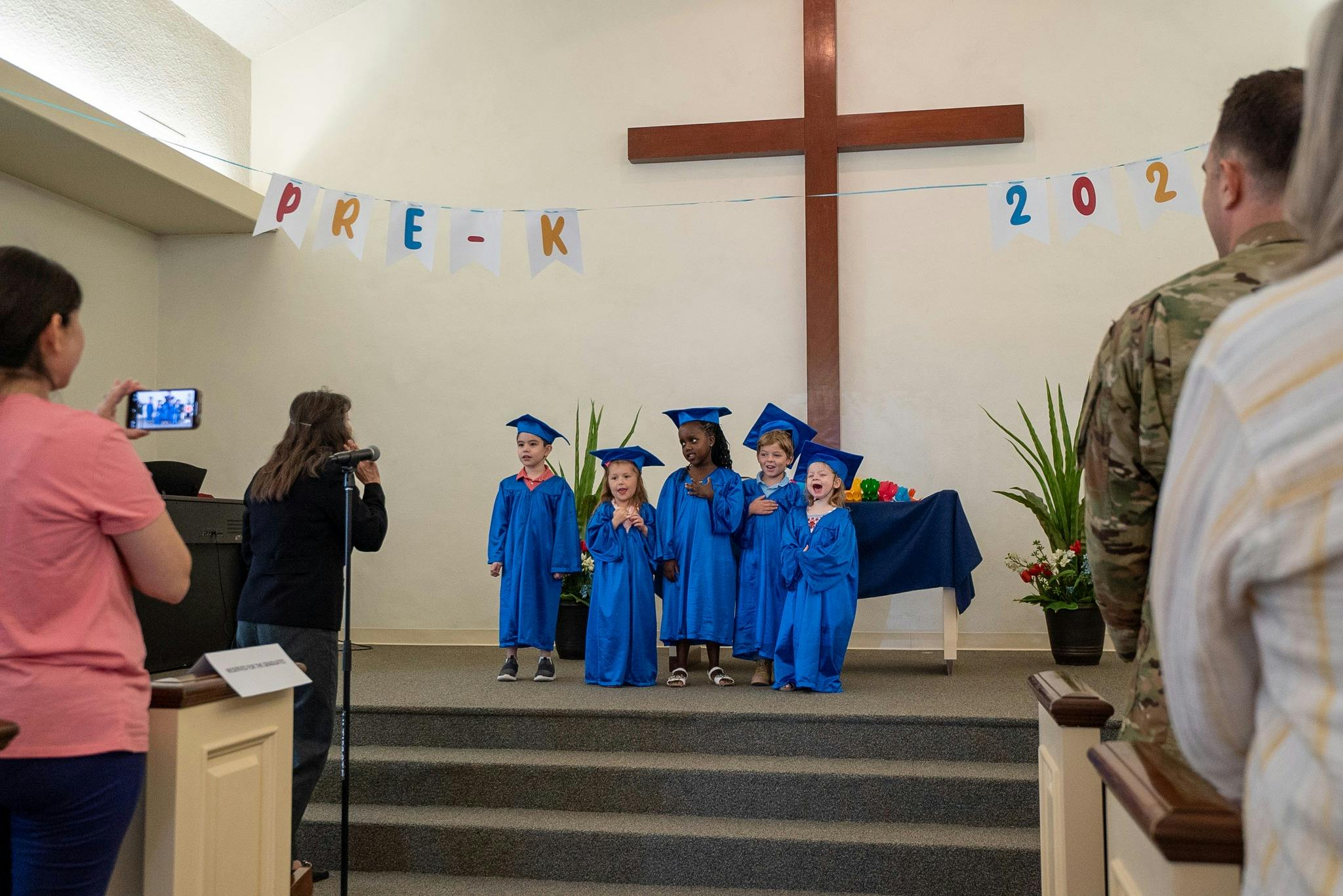 Children in blue graduation gowns and caps on a stage with a cross behind them