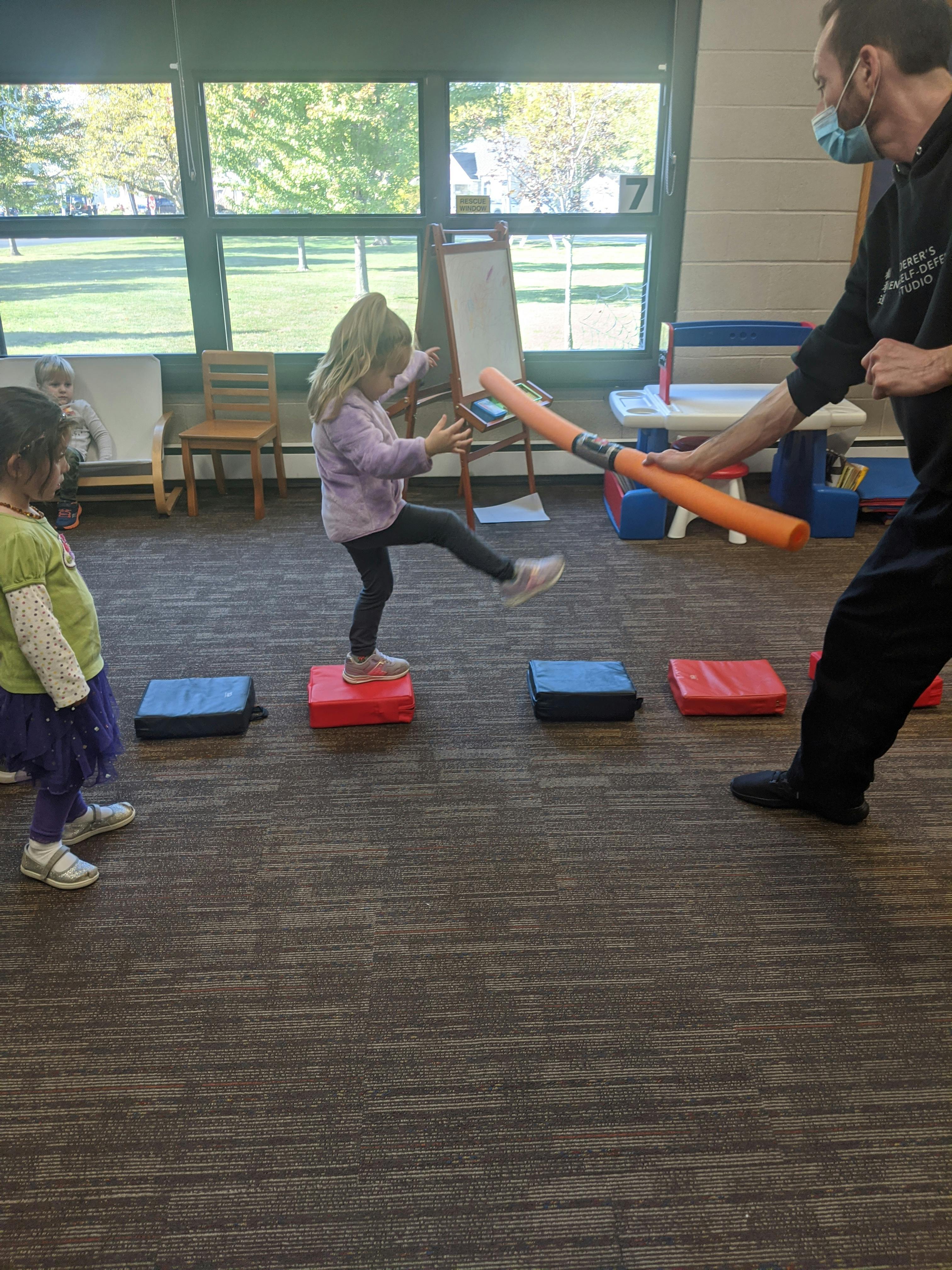 Child balancing on a red mat while a person holds an orange foam tube