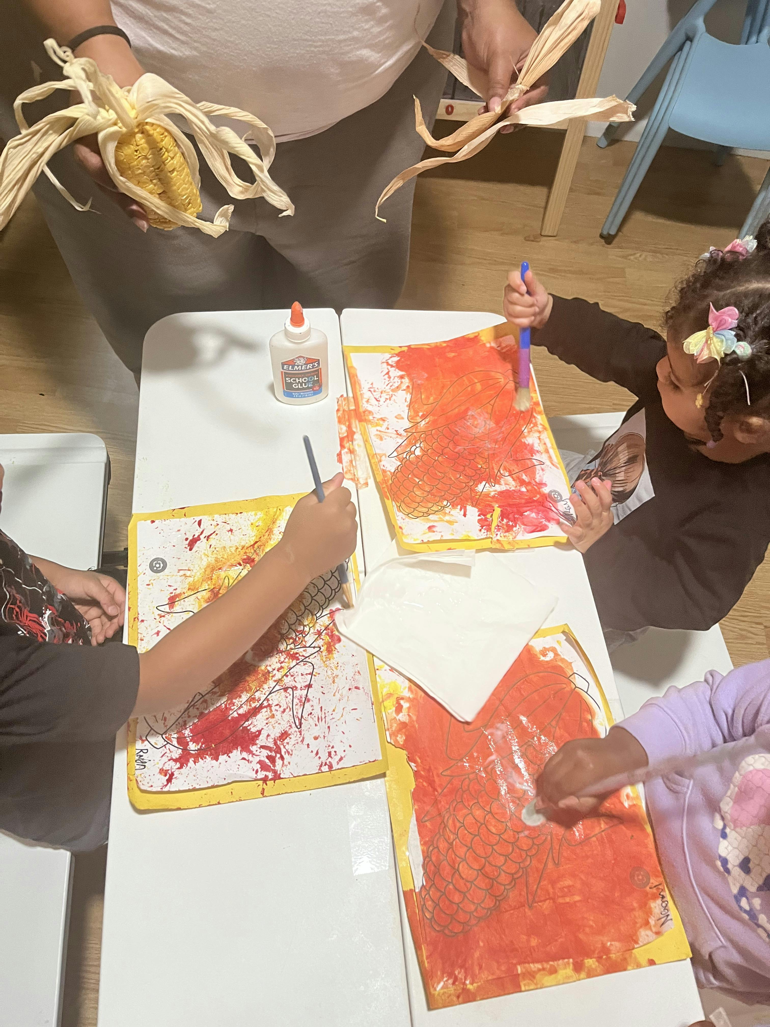 Children painting at a table, one adult holding corn with husks