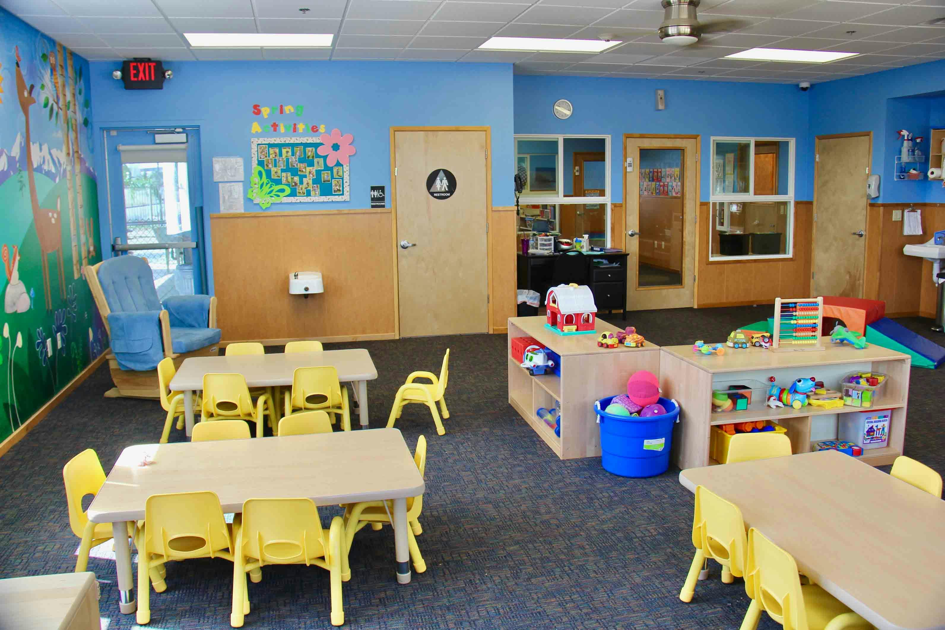 Empty classroom with small tables and chairs, and shelves with toys