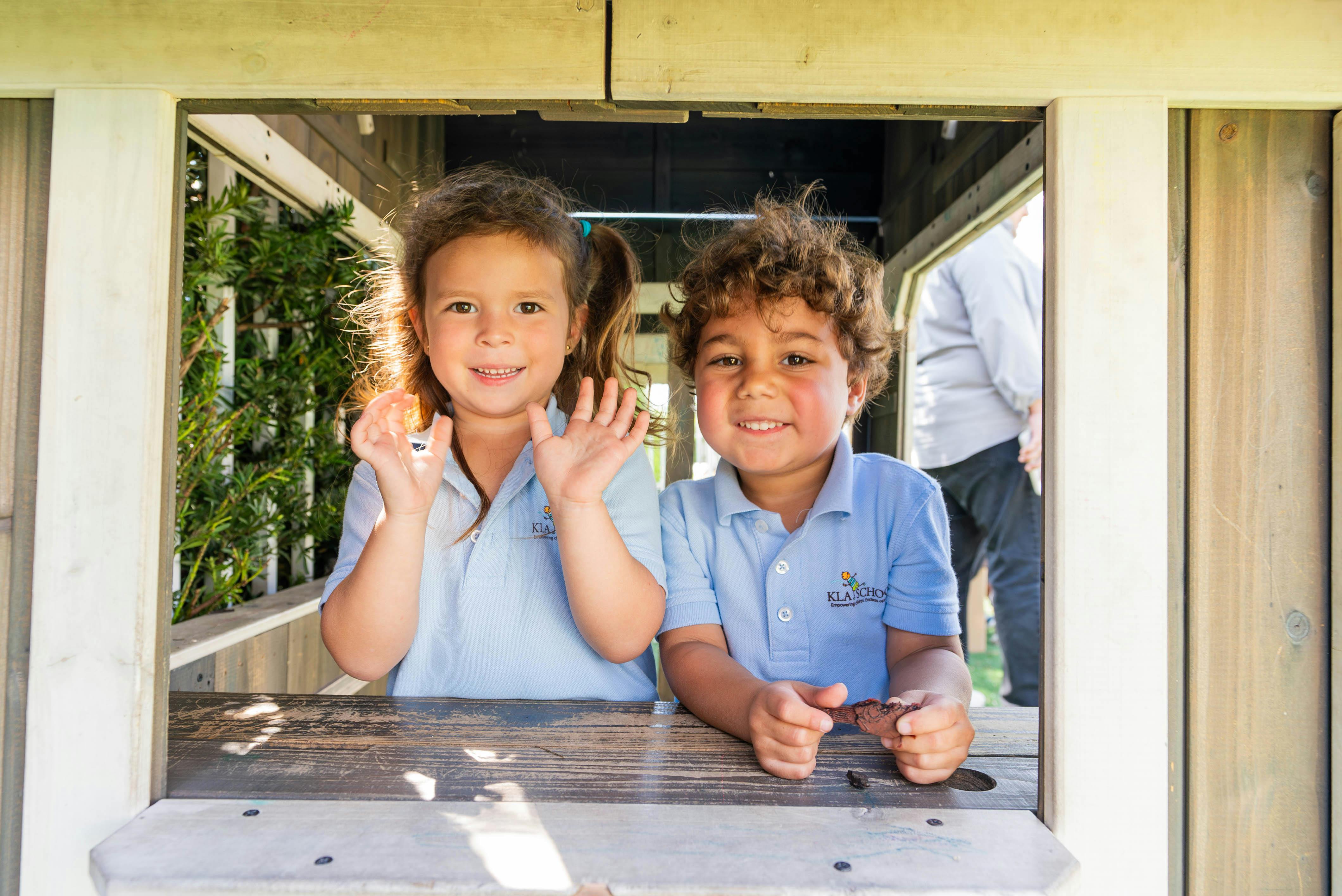 Two children smiling and looking out a window from a wooden playhouse