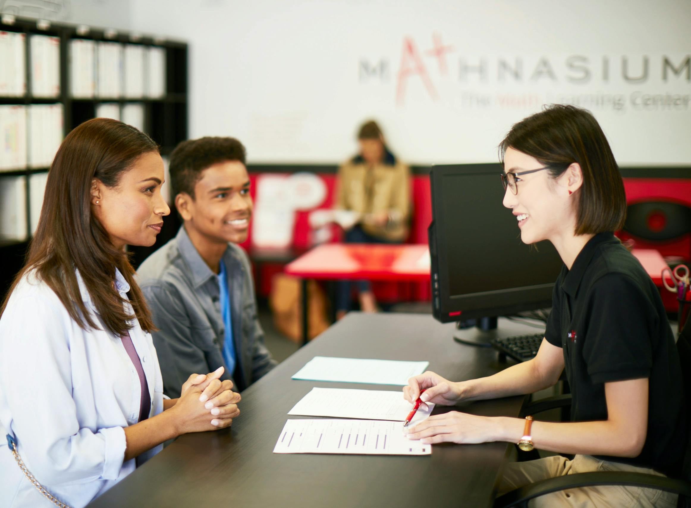 Three people at a desk, one writing on papers, two observing