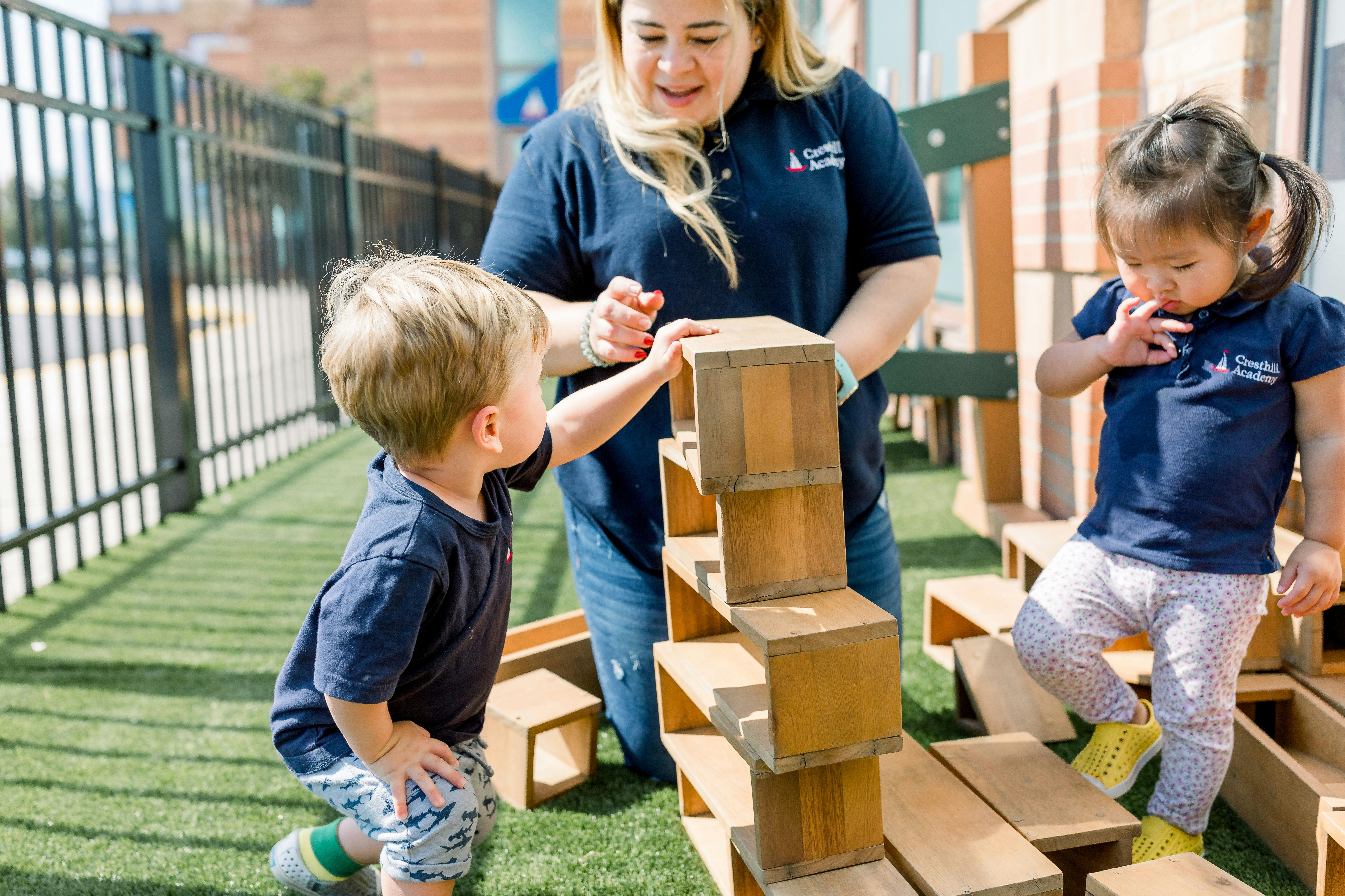 Adult and two children playing with large wooden blocks on artificial grass