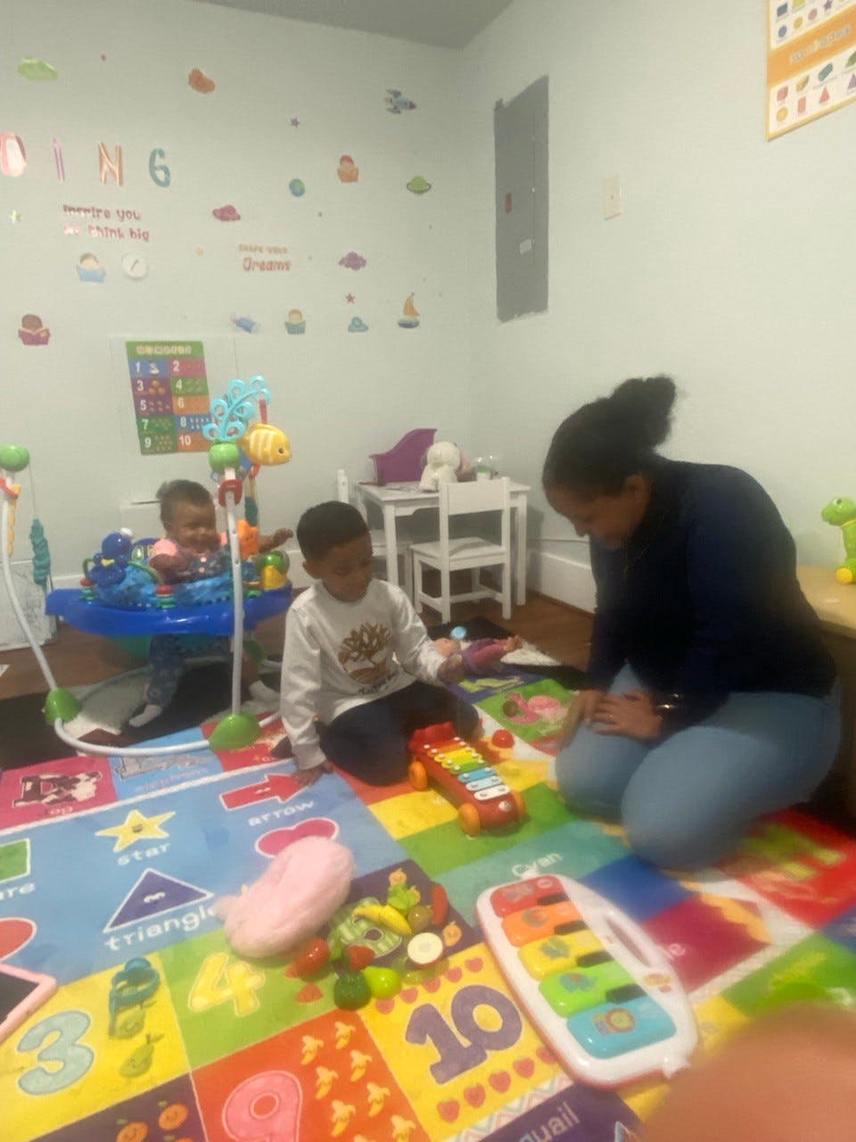 Adult and child playing with toys on a colorful mat, another child in a bouncer