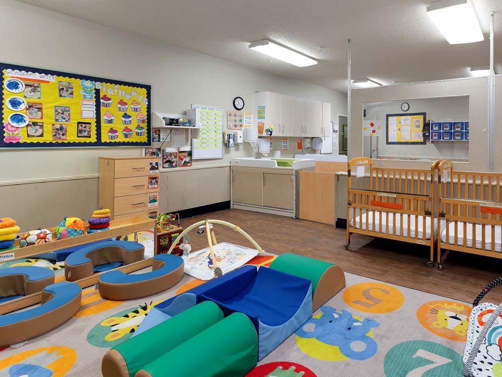 A colorful daycare room with play mats, wooden cribs, and a bulletin board