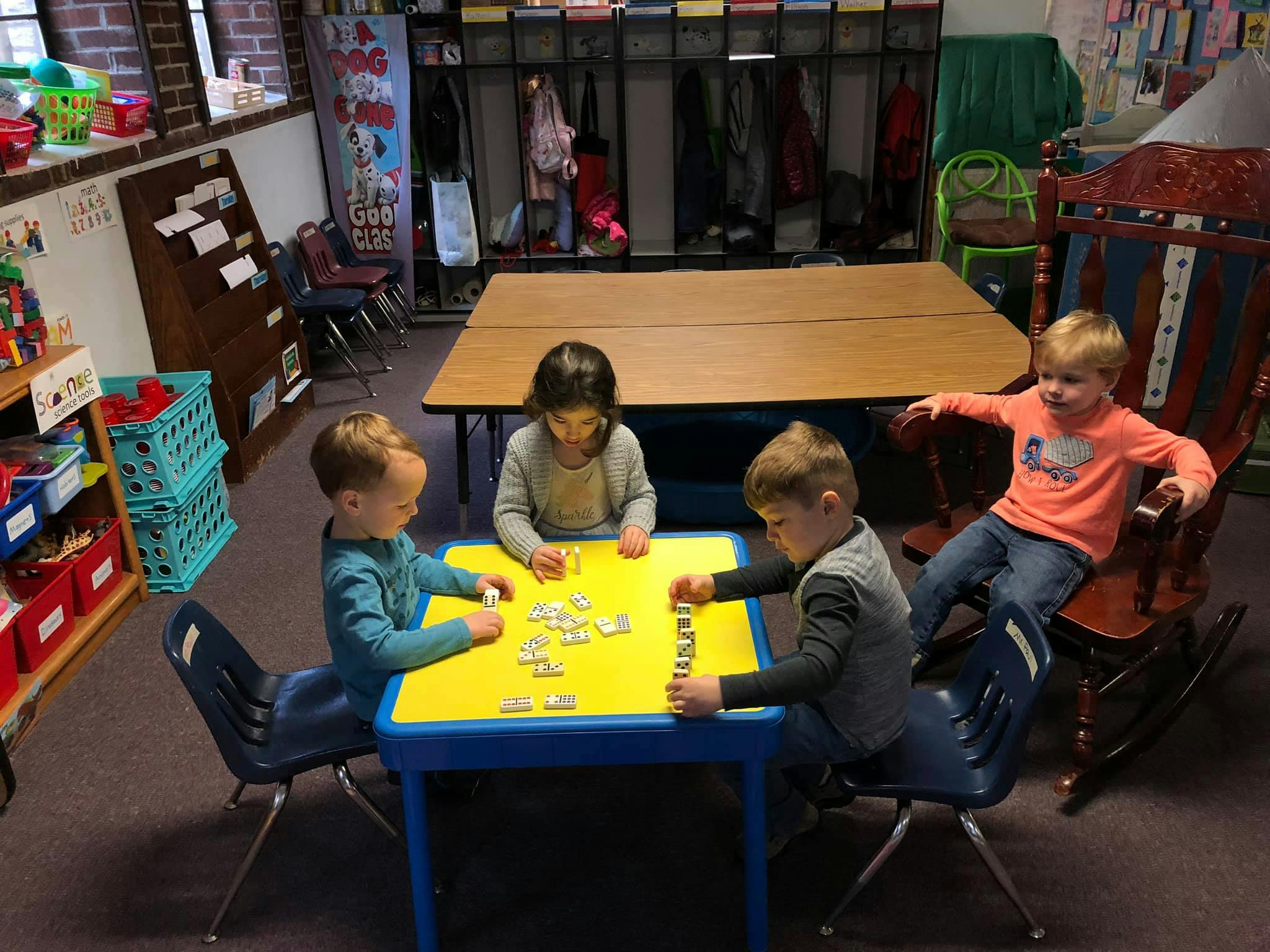Children playing with dominoes at a yellow table in a classroom