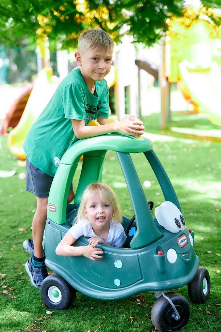 Child standing behind a toy car with a younger child sitting inside