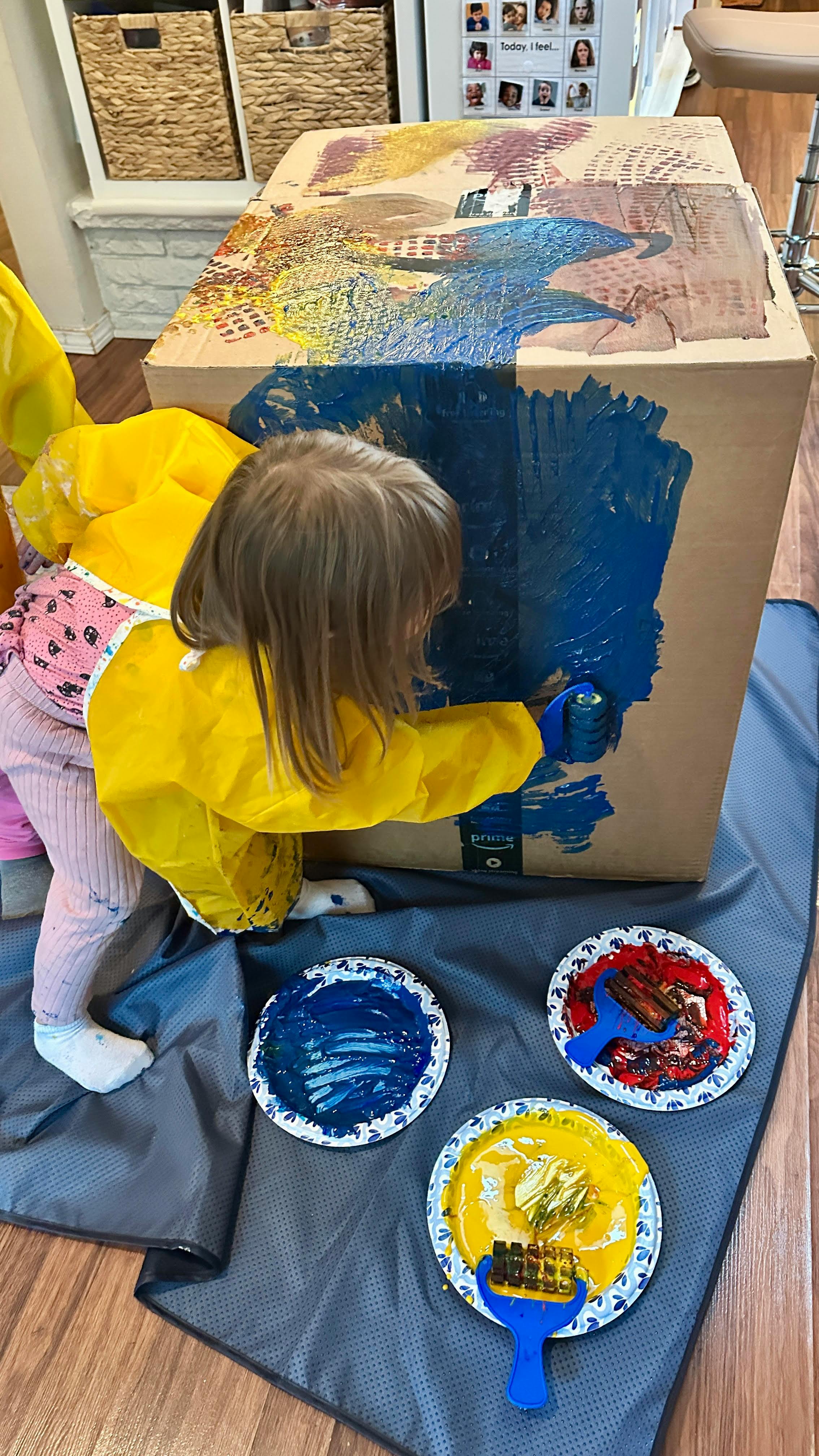 Child in yellow smock painting a large cardboard box blue