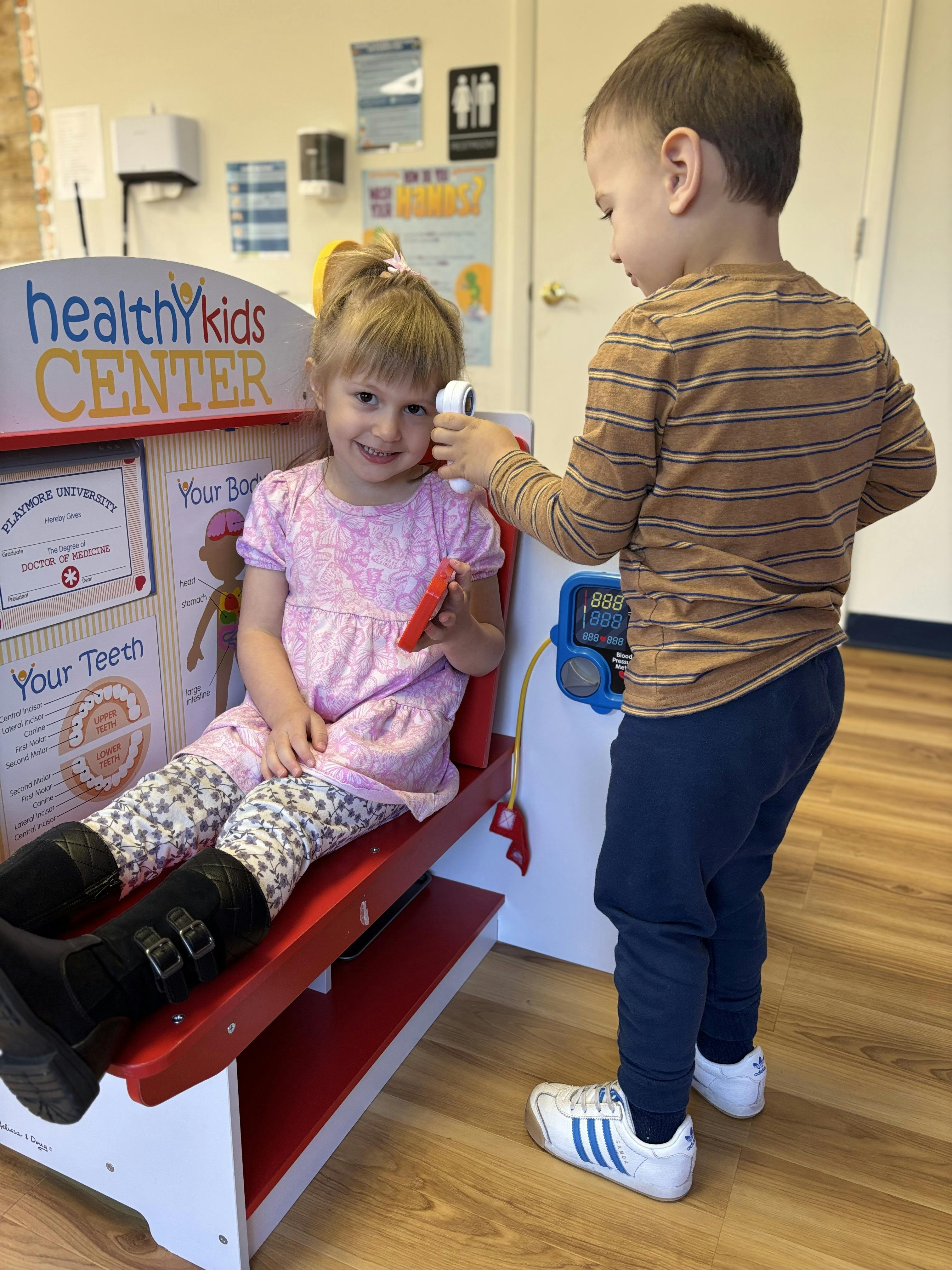 Child sitting in a play doctor's office while another child checks their temperature