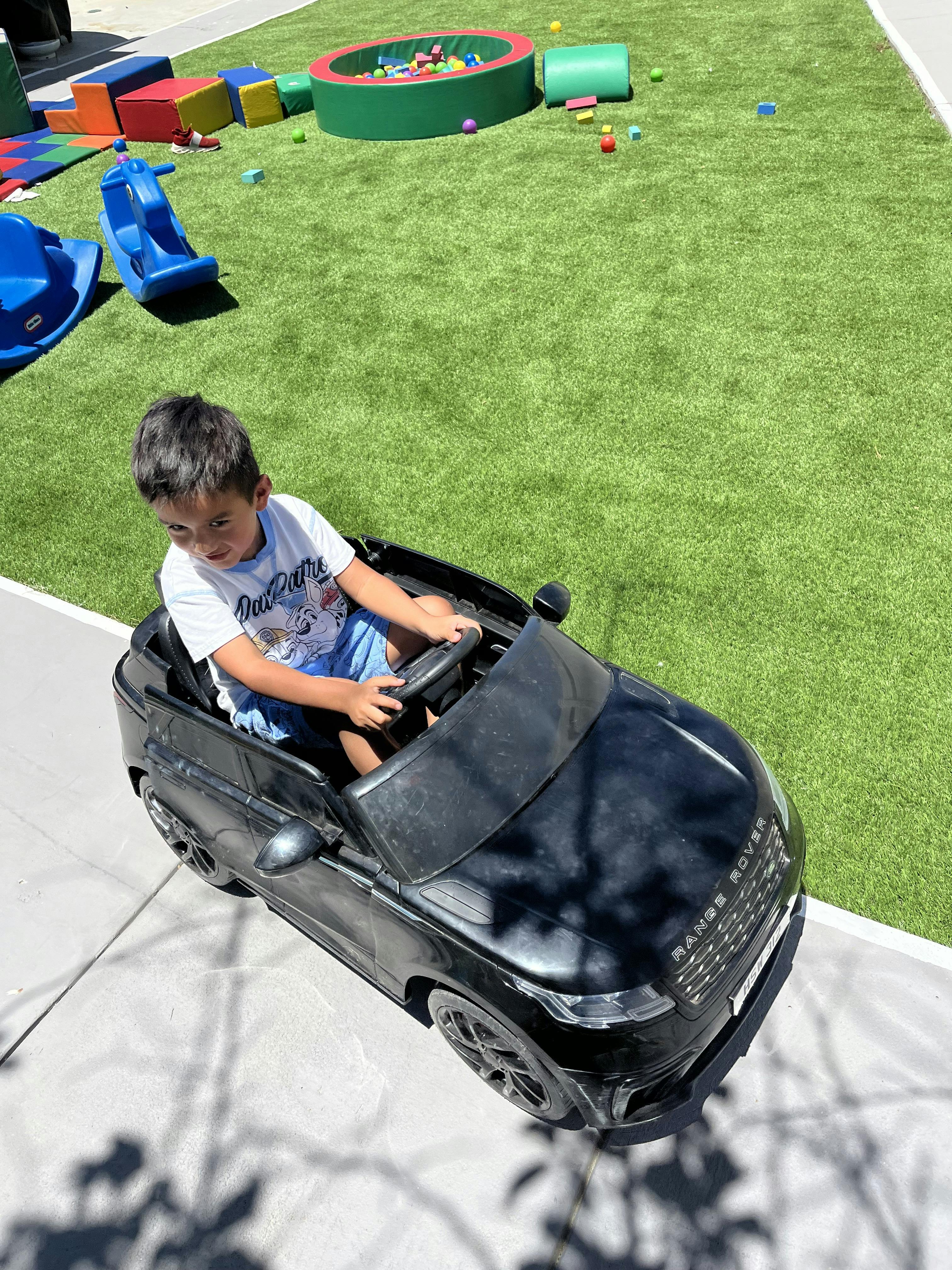 Child in a toy car on a sidewalk next to a grassy area with toys