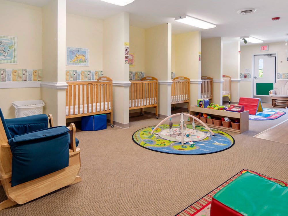 A nursery room with multiple cribs, a blue rocking chair, and playmats with toys
