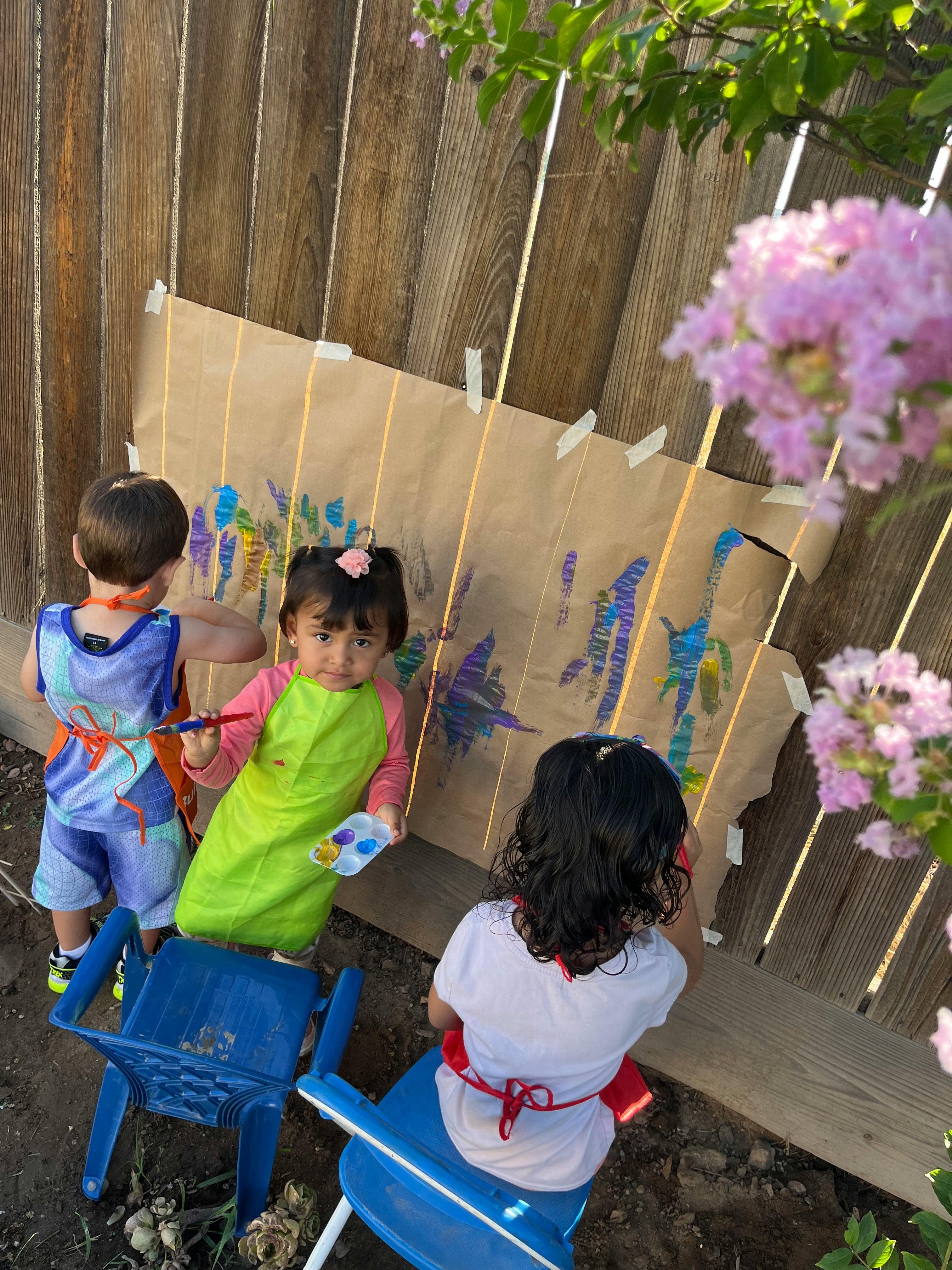 Three children painting on a large piece of paper taped to a wooden fence