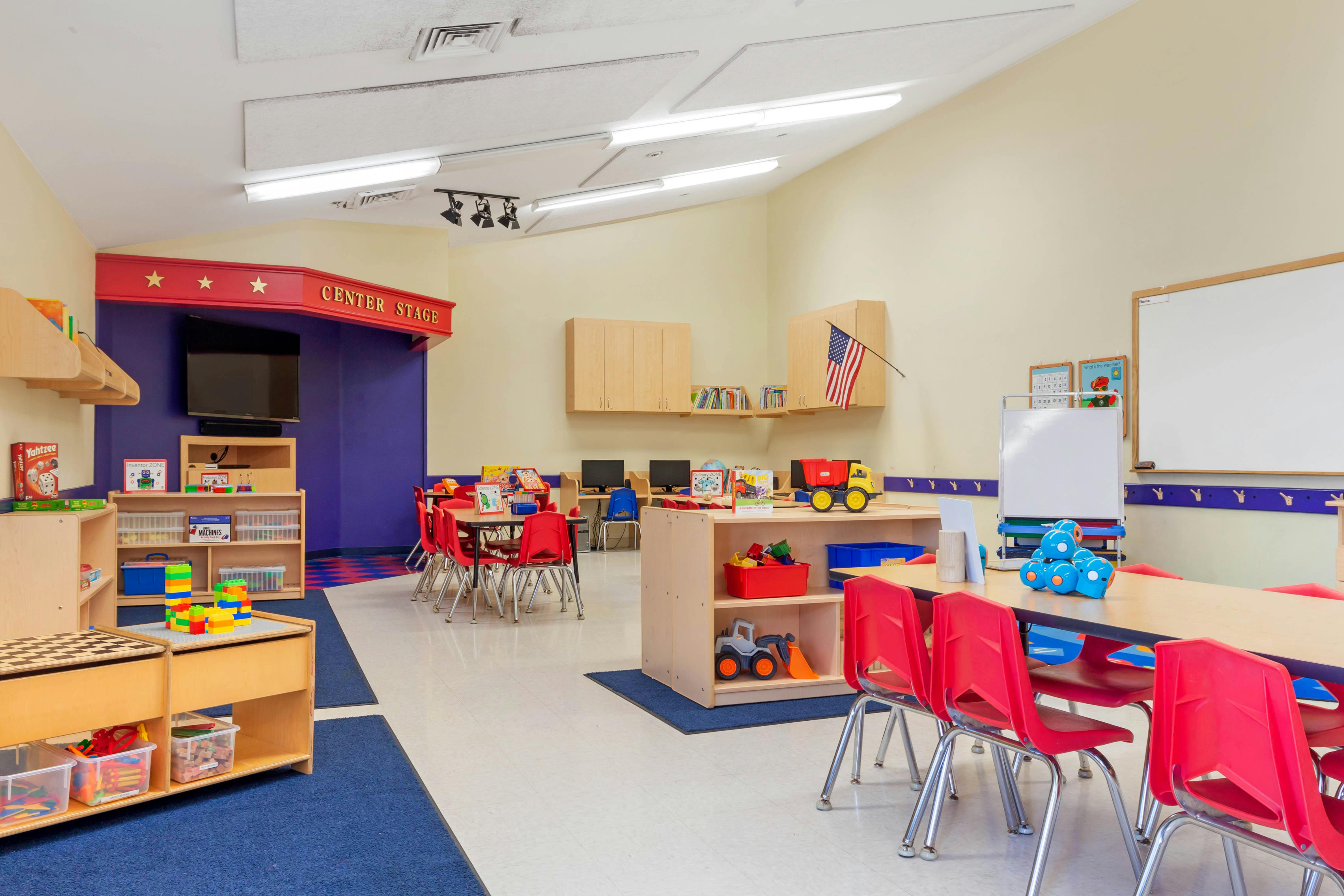 A classroom with small tables and chairs, shelves with toys, and a "Center Stage" area