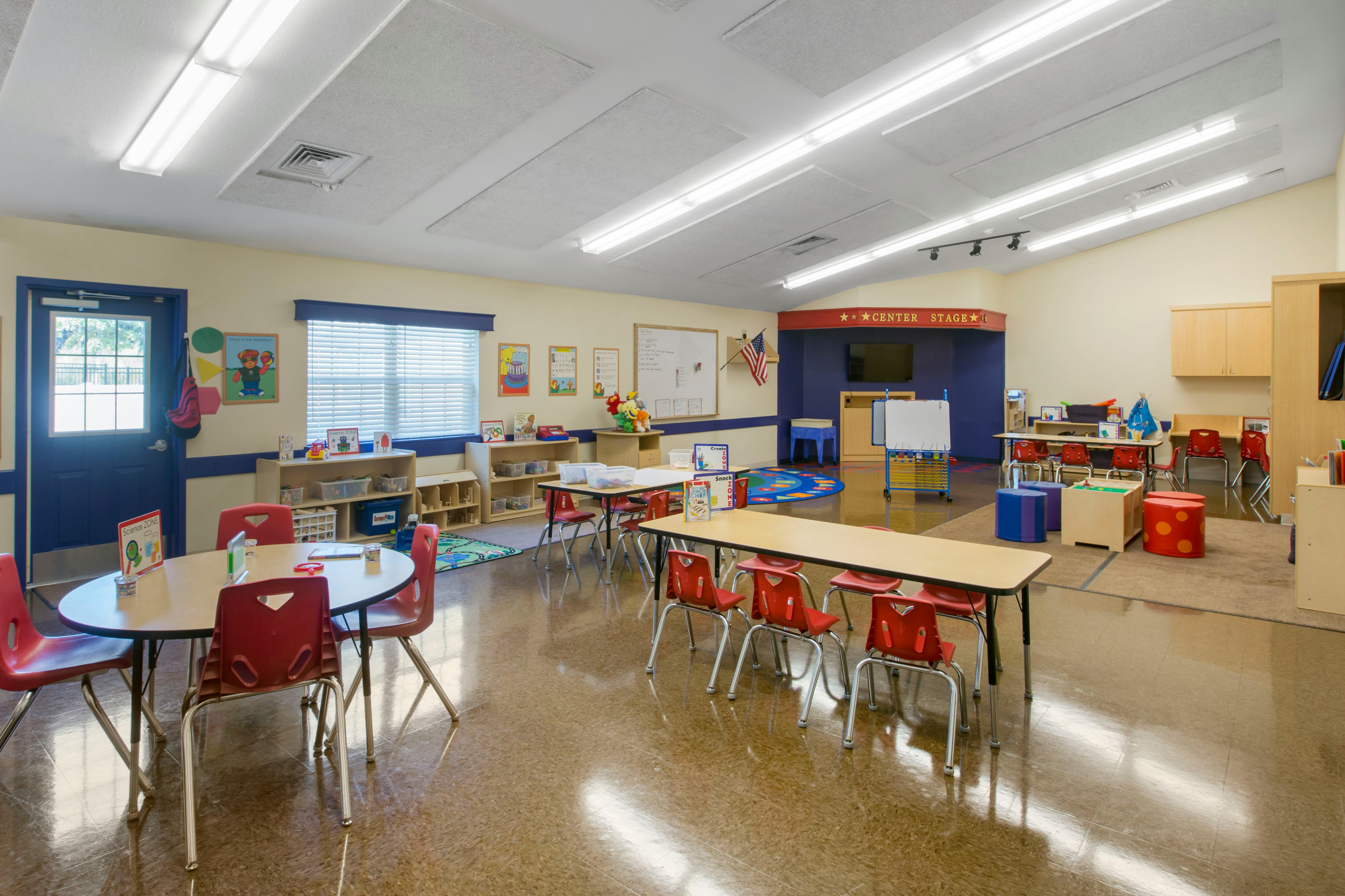 Classroom with small tables and red chairs, display board, whiteboards, and play areas