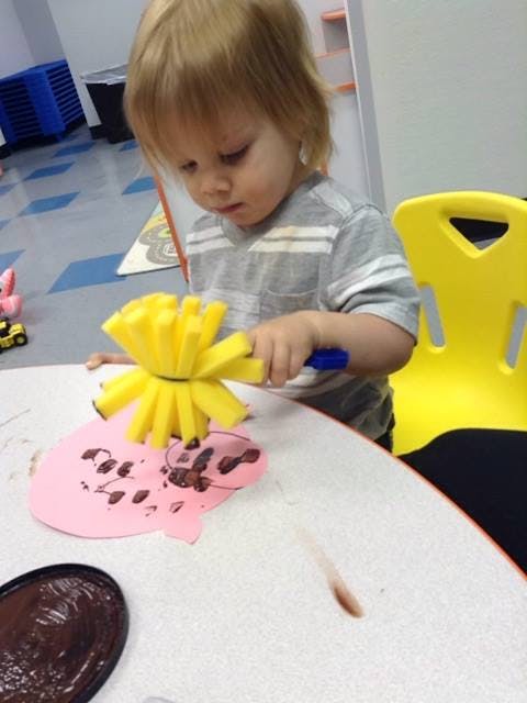 Child using a yellow sponge brush to paint a pink paper cutout
