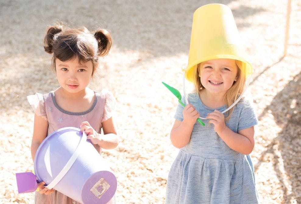 Two children playing outdoors, one holding a bucket, one with a bucket on head