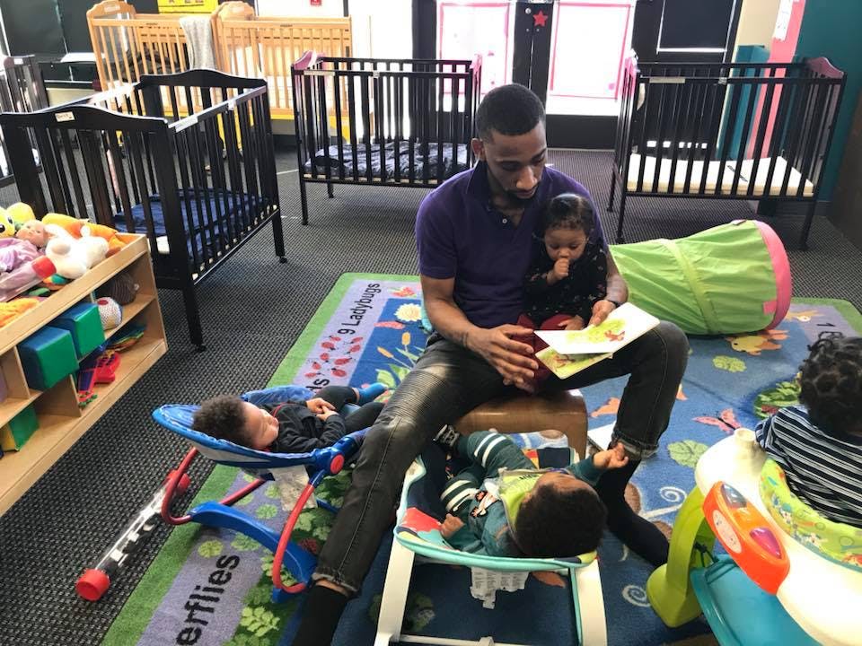 Person holding a child and reading a book to children in a room with cribs