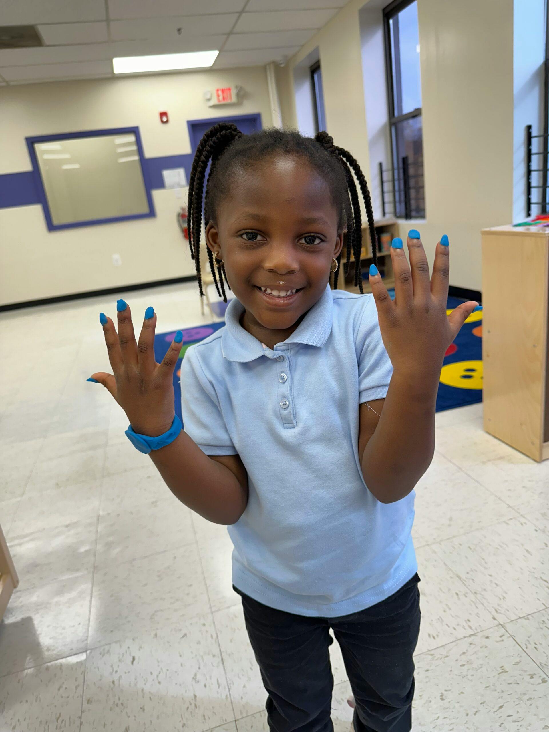Child in a light blue shirt showing fingers with blue play-doh on the nails