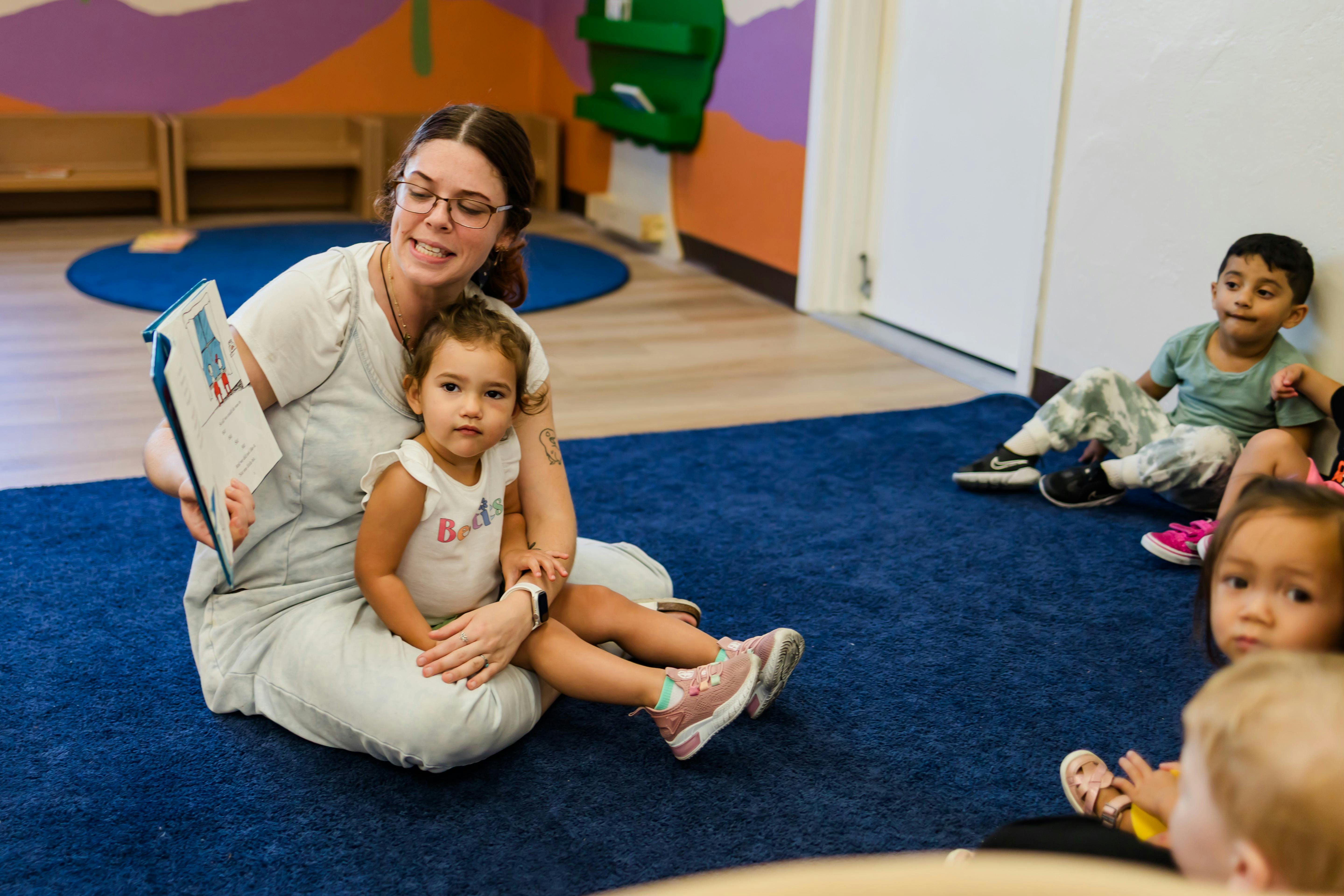 Person reading a book to a child on their lap, other children nearby