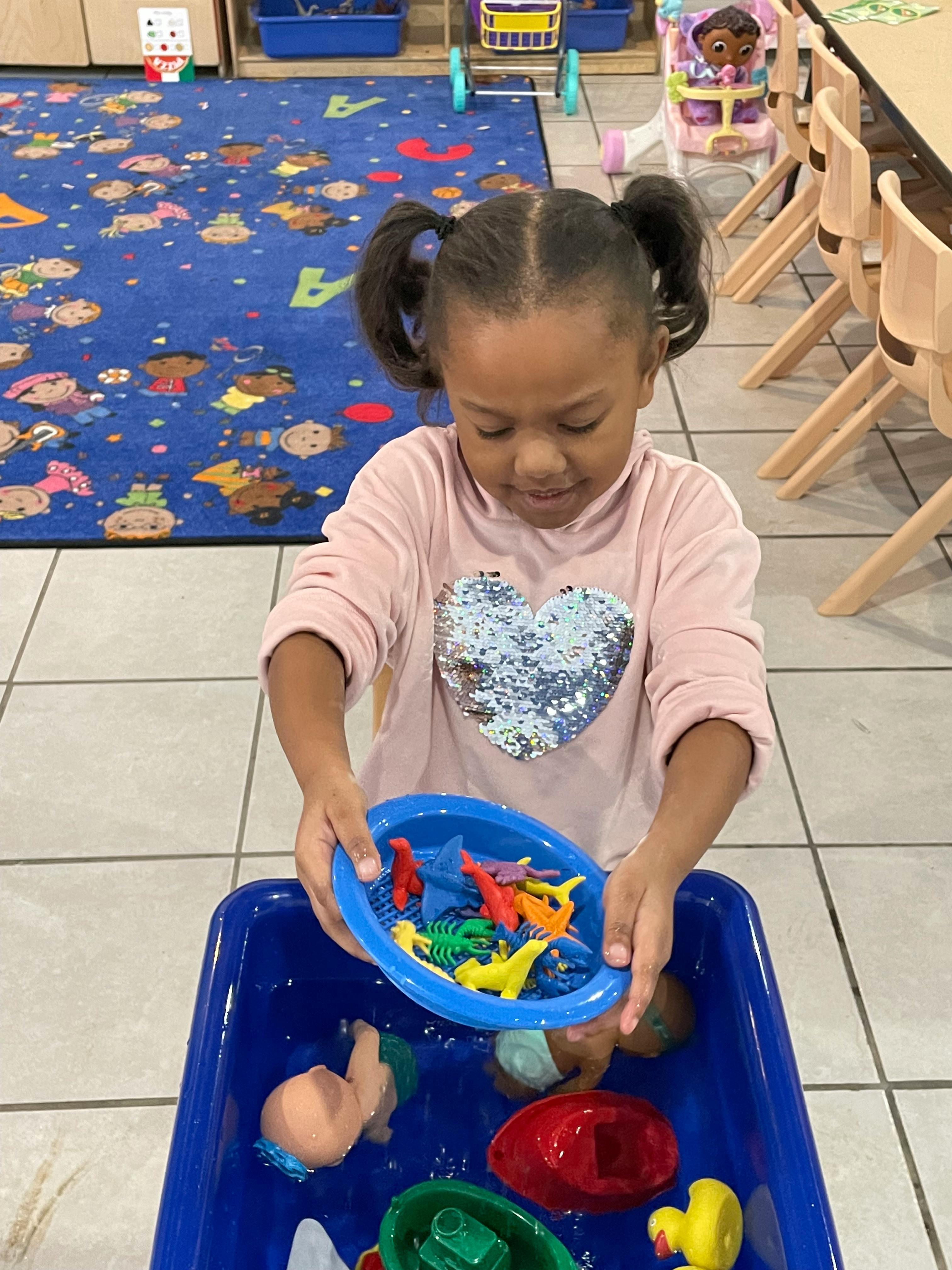 Child holding a blue bowl of toy animals over a blue bin with water