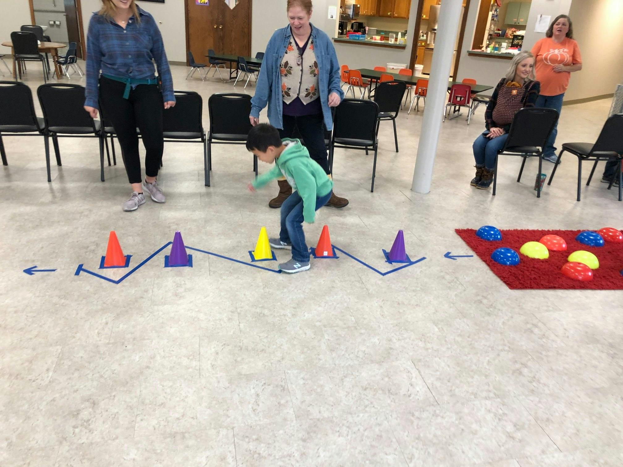 Child navigating an obstacle course with cones and a red mat, with adults watching