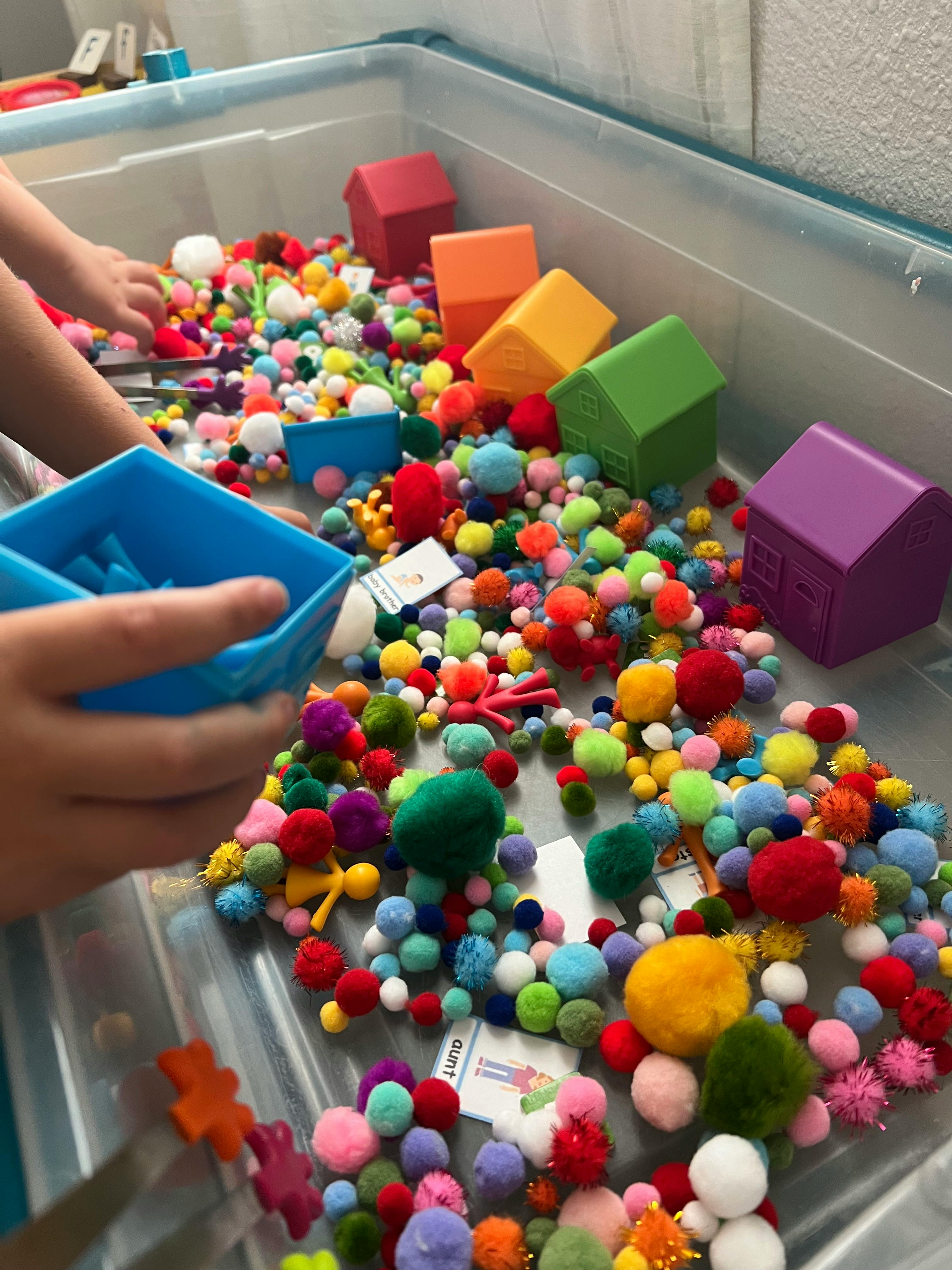 Child playing with colorful pom-poms and toy houses in a sensory bin
