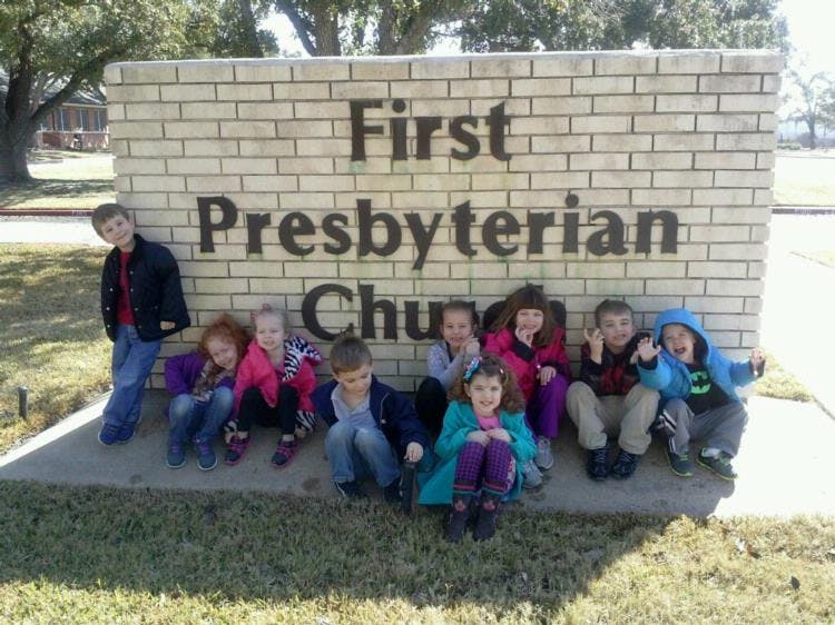 Children sitting and standing in front of a brick sign that reads "First Presbyterian Church"