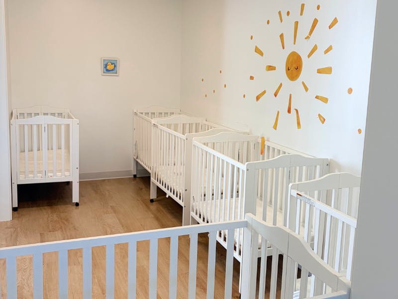 Five white cribs in a bright room with a sun decal on the wall