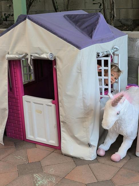 Child looking out the window of a toy playhouse with a stuffed unicorn