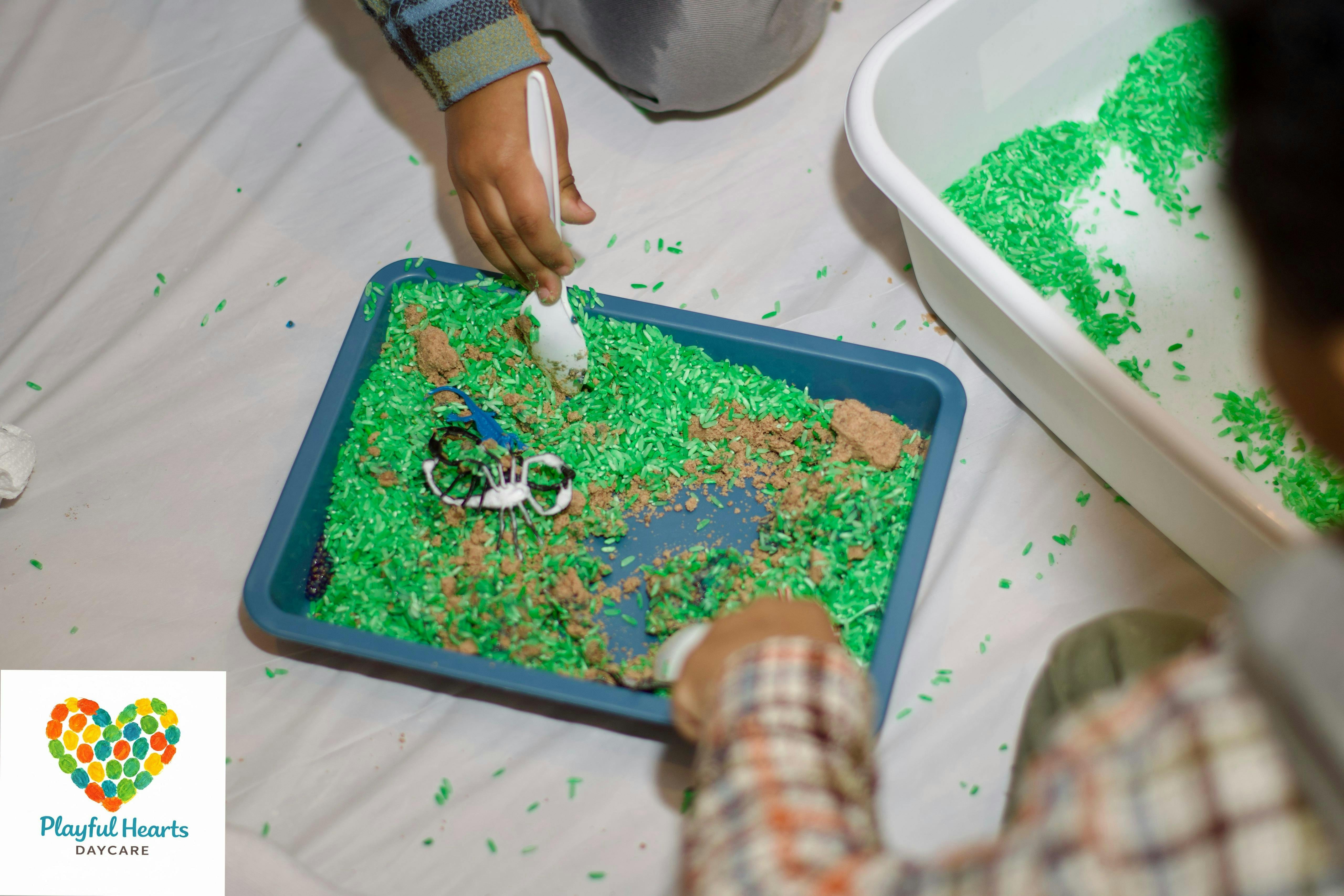 Child's hands playing with green rice, toy insects, and brown sand in a sensory bin