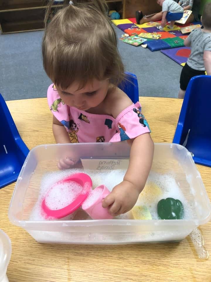 Child playing with soapy water and pink cups in a plastic bin