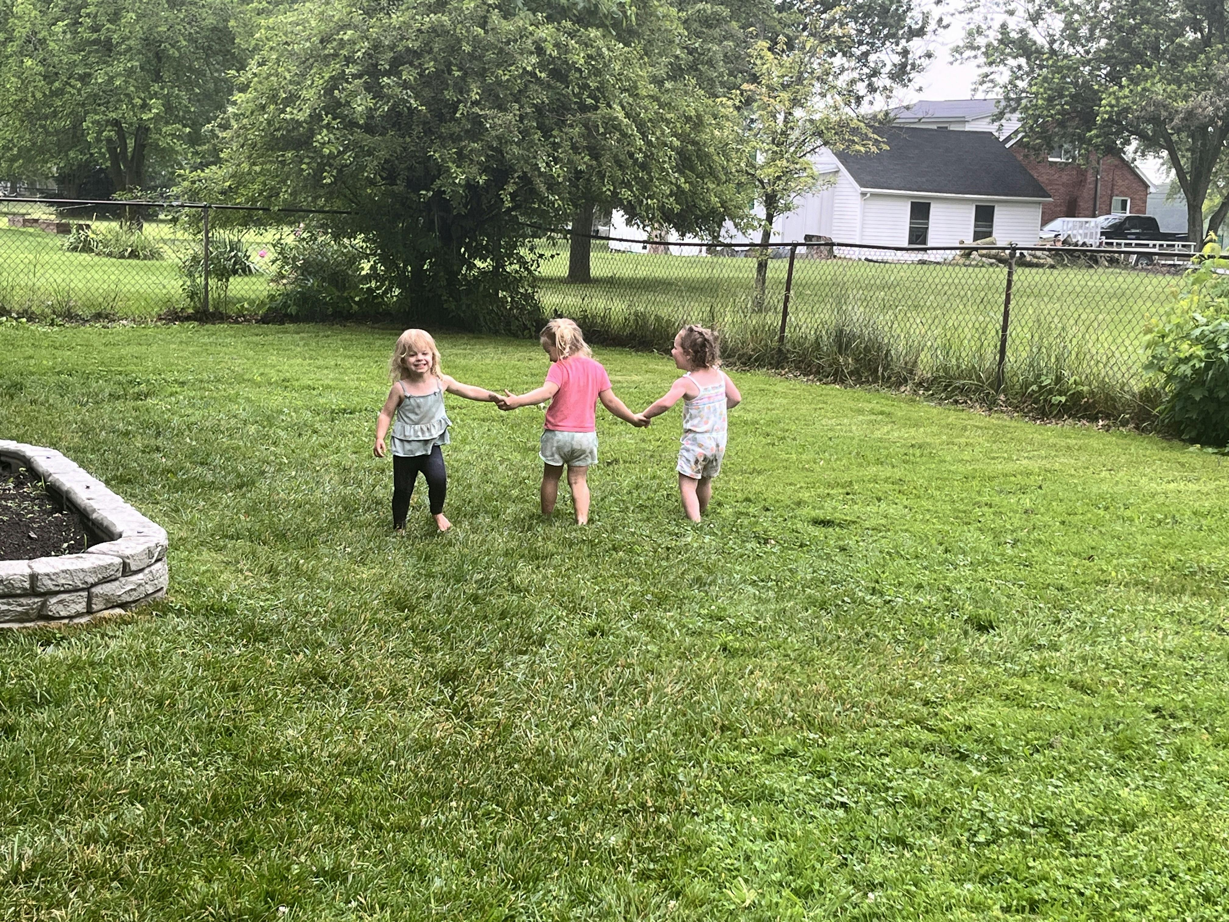 Three children holding hands walking in a grassy yard