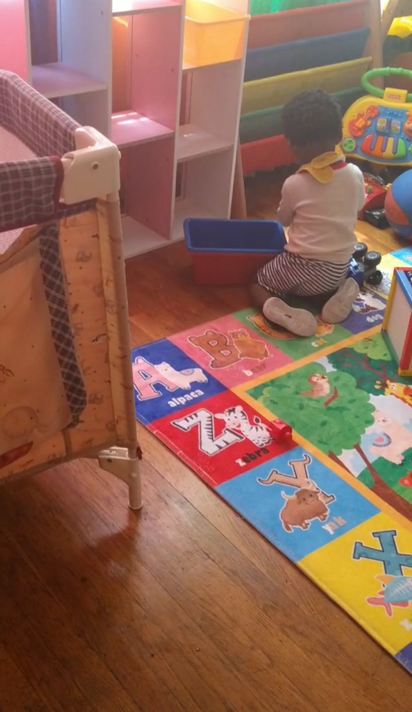Child sitting on an alphabet playmat in a room with a crib and shelves