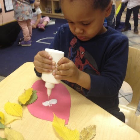 Child squeezing glue onto a pink heart-shaped paper with leaves on a table