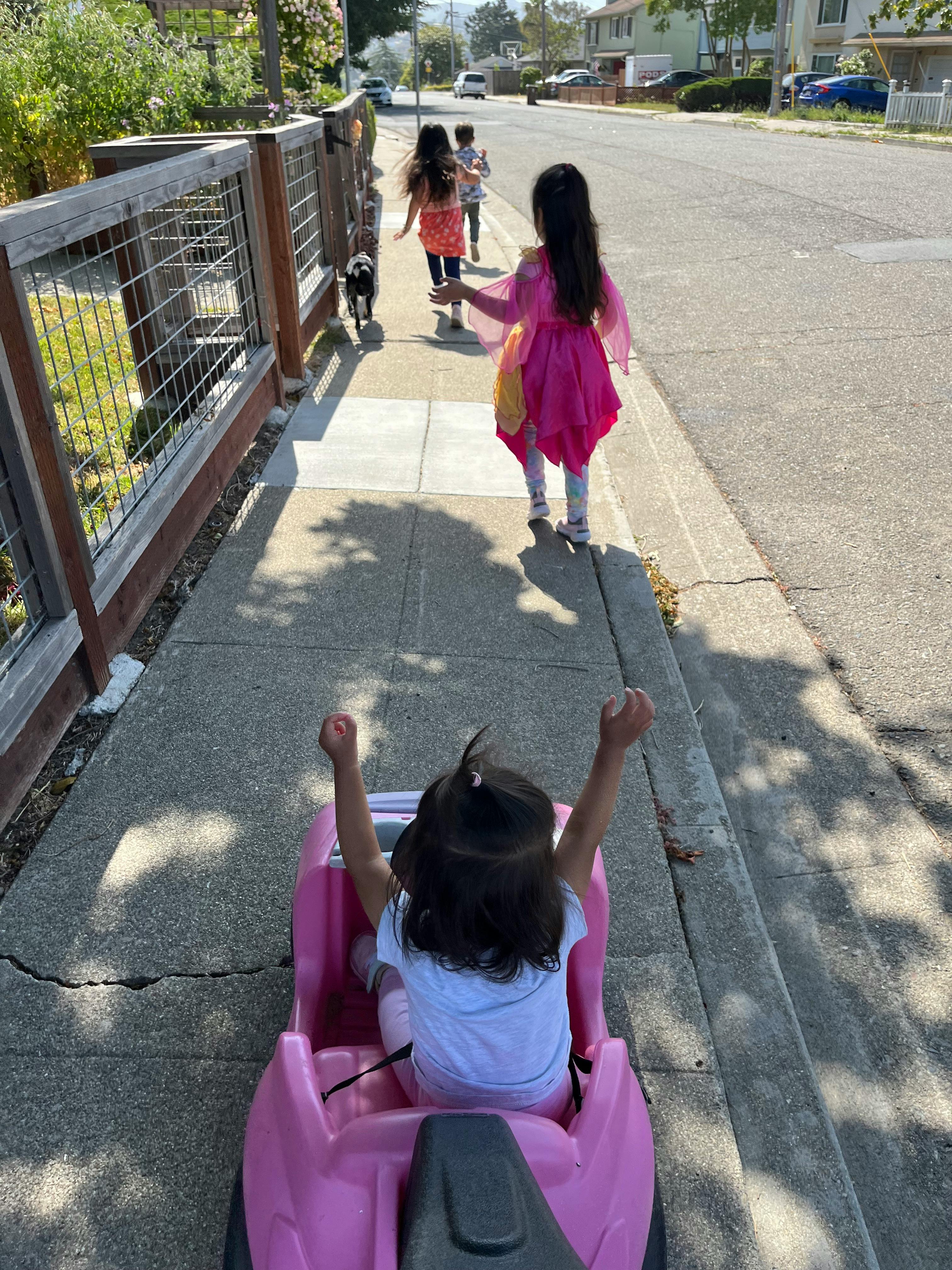 Child in pink toy car with arms raised, following other children on sidewalk
