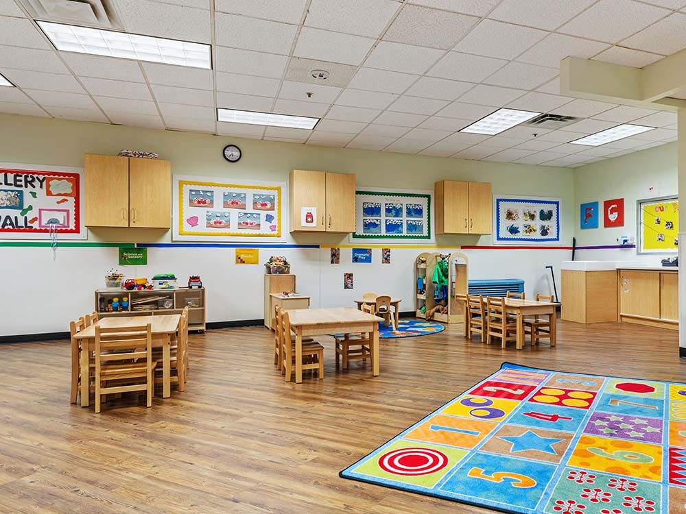 empty preschool classroom with small tables and chairs, and colorful rug